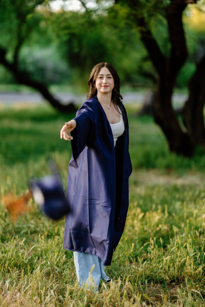 Senior photo throwing cap and wearing gown in the green grass and trees at Butcher Jones Recreation Site, Salt River, Mesa, Arizona.