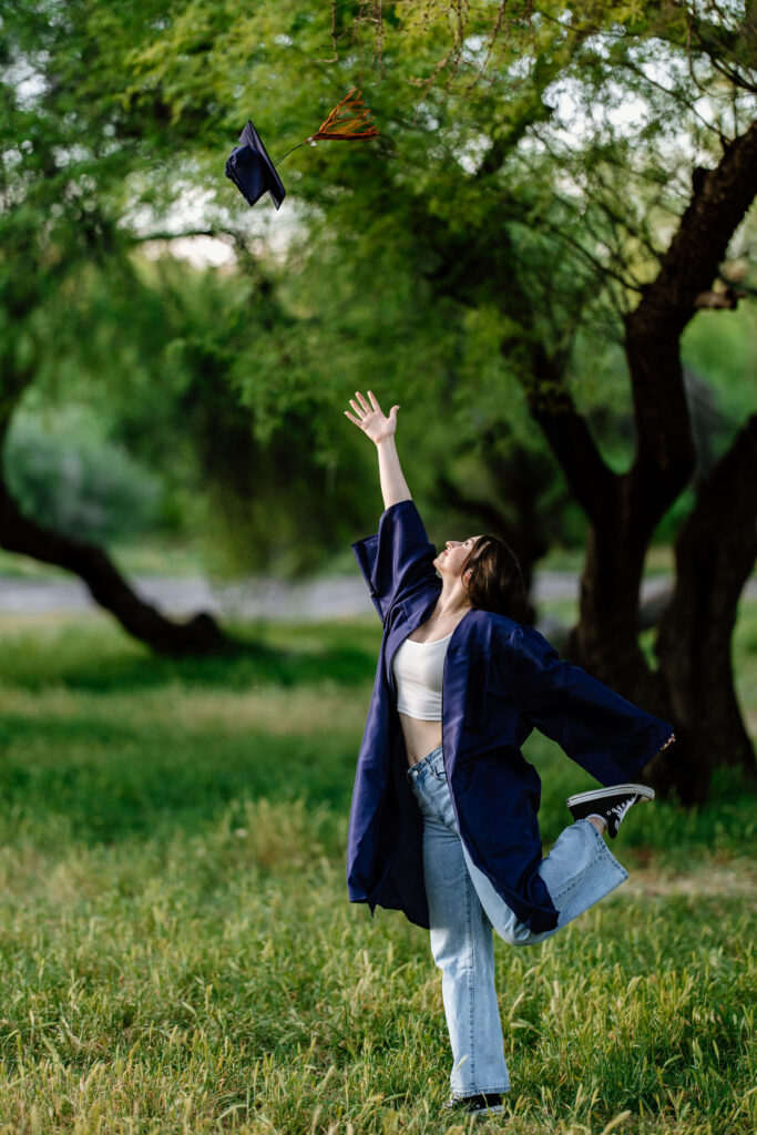 Senior photo throwing cap and wearing gown in the green grass and trees at Butcher Jones Recreation Site, Salt River, Mesa, Arizona.